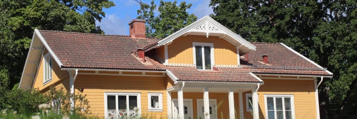 A picturesque yellow bungalow with a tiled roof surrounded by lush greenery under a bright blue sky.